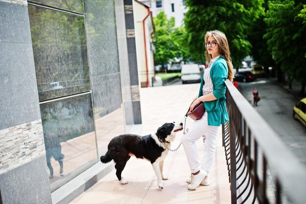 Trendiges Mädchen mit Brille und zerrissenen Jeans mit russoeuropäischem Laika Husky-Hund an der Leine gegen Gebäude auf der Straße der Stadt Freund Mensch mit Tierthema