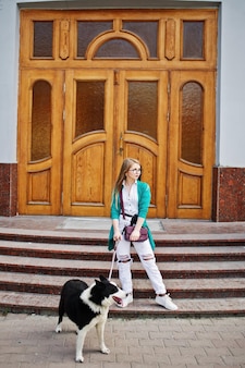 Trendiges mädchen mit brille und zerrissenen jeans mit russoeuropäischem laika husky-hund an der leine gegen holztür auf der straße der stadt freund mensch mit tierthema