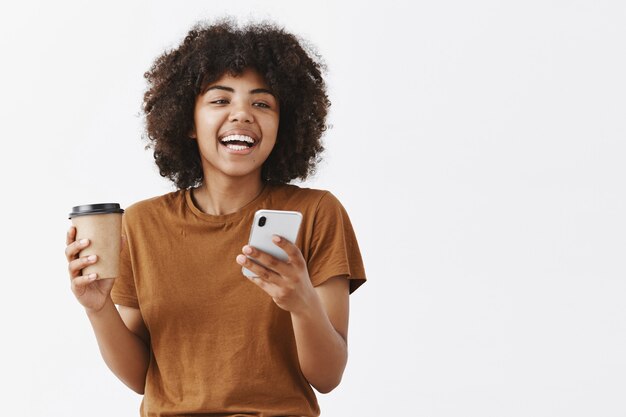 trendiges sorgloses Afroamerikanermädchen mit dem lockigen Haar im braunen T-Shirt lachend, während sie mit Freunden sprechen, die Kaffee vom Pappbecher trinken und Smartphone halten