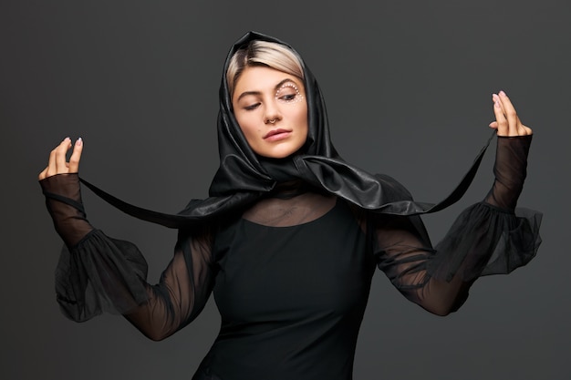 Trendy aussehende stilvolle junge Frau mit künstlerischem glamourösem Make-up posiert mit transparenter Bluse und schwarzem Lederkopftuch um den Hals. Schönheits- und Modekonzept