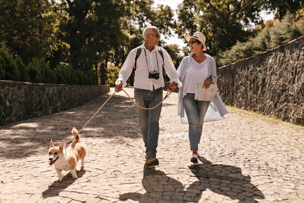 Trendy Frau im Hut und in der blauen Bluse, die grauhaarigen Mann im weißen Langarmhemd mit Kamera und Corgi im Park gehen und suchen.