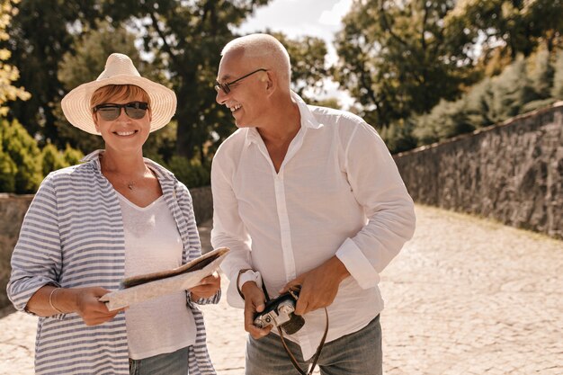 Trendy Frau mit kurzen Haaren im Hut, schwarzer Sonnenbrille und gestreifter Bluse lächelnd, Karte haltend und posierend mit grauhaarigem Mann mit Kamera im Park.