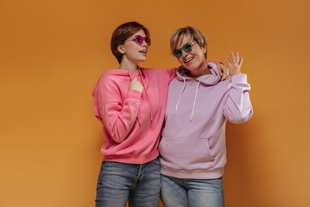 Trendy Frauen mit kurzen Haaren und hellen Sonnenbrillen in buntem Sweatshirt und kühlen Jeans, die auf orange lokalisiertem Hintergrund lächeln.