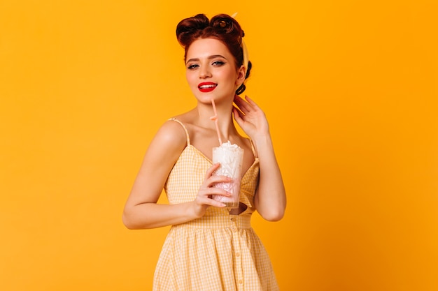 Trendy Ingwer Mädchen trinken Milchshake. Studioaufnahme von fit pinup Dame lokalisiert auf gelbem Raum.