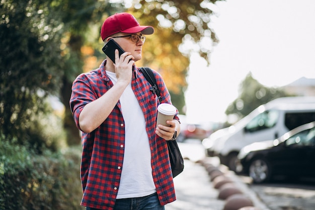 Trinkender Kaffee des jungen männlichen Studenten unter Verwendung des Telefons im Park