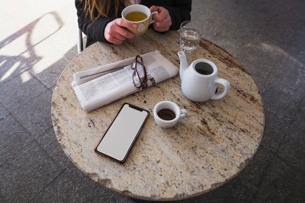 Trinkender Tee der Frau bei Tisch mit Zeitung und Smartphone