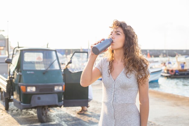 Trinkwasser der jungen Frau von der Flasche, die nahe der Küste steht