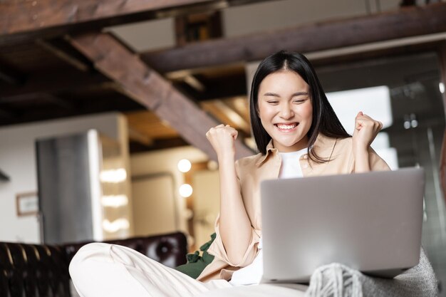 Triumphierendes asiatisches Mädchen, das online gewinnt und den Sieg feiert, während es sich zu Hause ein Video auf dem Laptop ansieht