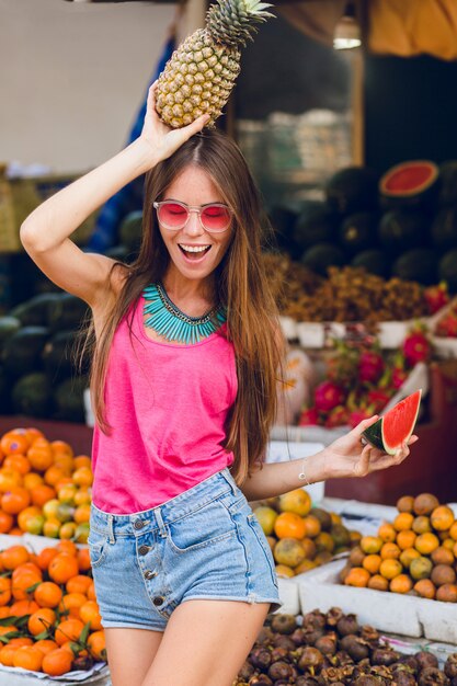 Tropisches Sommermädchen in der rosa Sonnenbrille auf dem Markt auf Obstmarkt. Sie hält Ananas auf dem Kopf und eine Scheibe Wassermelone. Sie sieht genossen aus