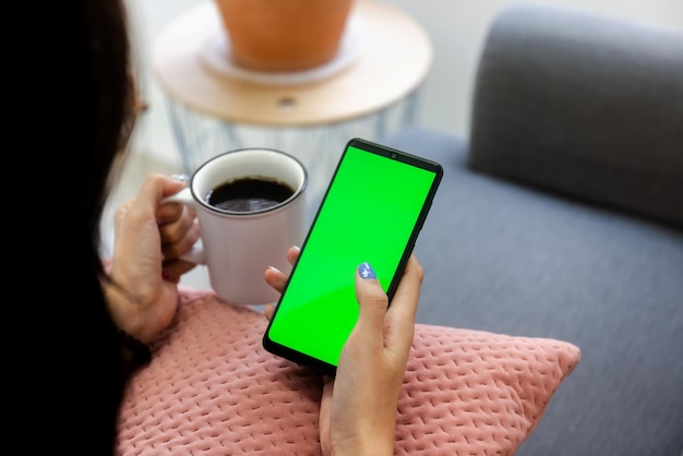 Über die schulter mädchen mit mobilem smartphone-computer auf dem sofa im wohnzimmer mit leerem grünem bildschirm arbeiten von zu hause aus konzept