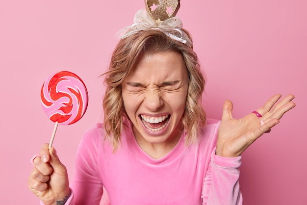 Kostenloses Foto Überglückliche frau ruft vor freude aus und hält handgehobene posen mit süßen lollipop-kleidern für partys mit optimistischen stimmungsposen vor rosa hintergrund. fröhliches mädchen im festlichen outfit hält karamellbonbons