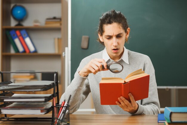 überrascht aussehendes Buch mit Lupe junger männlicher Lehrer sitzt am Schreibtisch mit Schulwerkzeugen im Klassenzimmer