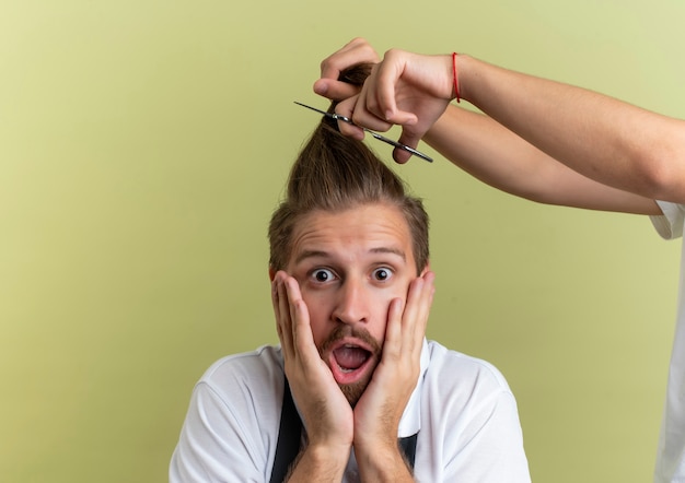 Überrascht junger hübscher friseur, der hände auf gesicht setzt, hat angst, alle seine haare abgeschnitten auf olivgrüner wand abgeschnitten zu bekommen