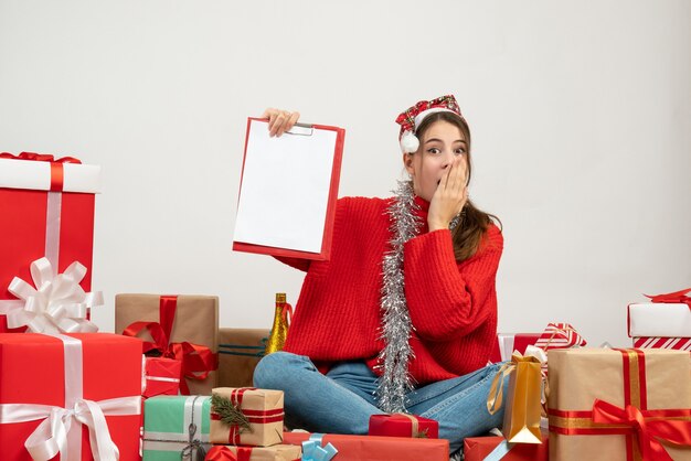 überrascht süßes Mädchen mit Weihnachtsmütze hält Dokument sitzen um Geschenke auf Weiß