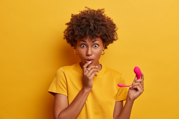 Kostenloses Foto Überraschte dunkelhäutige frau, die von innovativen technologien im sexbereich schockiert ist, einen intelligenten vibrator aus rosa gummi hält, der mit ihrer vorhandenen technologie synchronisiert werden kann, und über bluetooth eine verbindung zum mobiltelefon herstellt