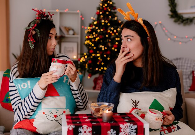 überraschte hübsche junge Mädchen mit Stechpalmenkranz und Rentierstirnband halten Tassen, die sich gegenseitig ansehen, auf Sesseln sitzen und die Weihnachtszeit zu Hause genießen