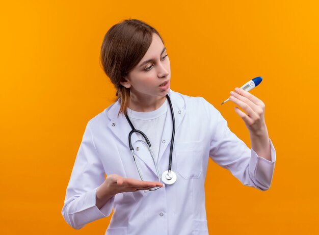 Überraschte junge Ärztin, die medizinische Robe und Stethoskop trägt und Thermometer hält, das es auf isolierter orange Wand betrachtet