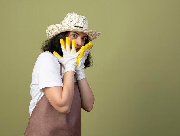 Kostenloses Foto Überraschte junge brünette gärtnerin in uniform, die gartenhut und handschuhe trägt, legt hände