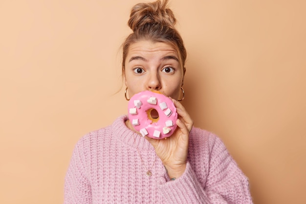 Kostenloses Foto Überraschte junge europäerin bedeckt den mund mit einem köstlichen, großen, appetitlichen donut, hat zuckersüchtiges haar, das in brötchen gekämmt ist, und trägt einen strickpullover, der über beigem hintergrund isoliert ist. naschkatzen-konzept