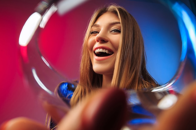 überraschte junge Frau in Partykleidung posiert mit Glas Wein. Emotionales weibliches süßes Gesicht. Blick aus dem Glas