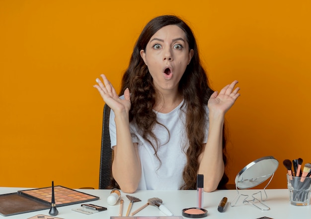 überraschte junge hübsche Frau, die am Make-up-Tisch mit Make-up-Werkzeugen sitzt, die leere Hände zeigen