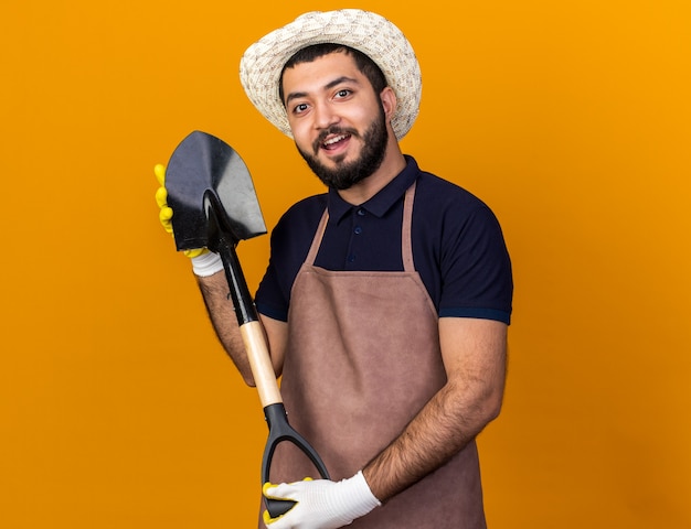 überraschte junge kaukasische männliche Gärtner, die Gartenhut und Handschuhe tragen, die Spaten lokalisiert auf orange Wand mit Kopienraum halten