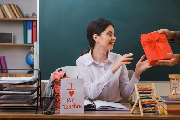 überraschte junge lehrerin erhielt eine geschenkbox, die mit schulwerkzeugen im klassenzimmer am schreibtisch saß