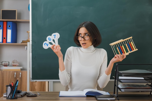 überraschte junge lehrerin mit brille, die abakus mit zahlenfächern hält, die am schreibtisch mit schulwerkzeugen im klassenzimmer sitzen