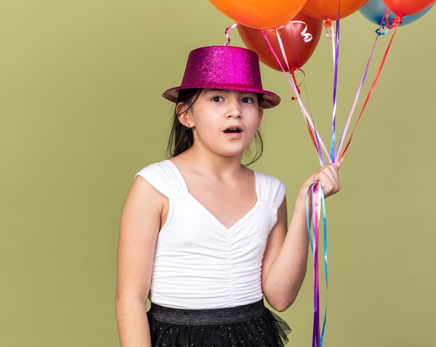 überraschte junges kaukasisches Mädchen mit lila Partyhut, der Heliumballons lokalisiert auf olivgrüner Wand mit Kopienraum hält