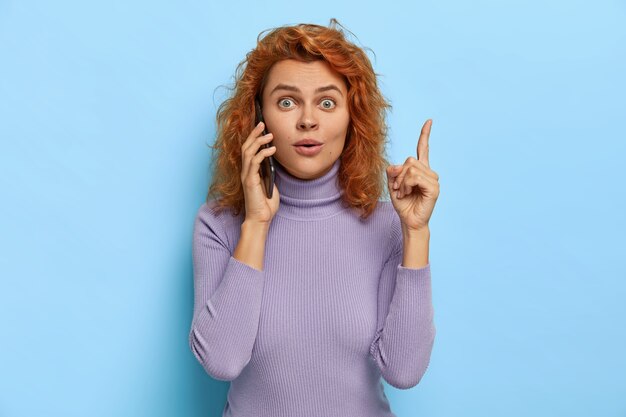 Überraschte schöne Ingwerdame bekommt eine ausgezeichnete Idee im Kopf, hebt den Zeigefinger, teilt ihren großartigen Plan mit einer Freundin über ein Smartphone-Gespräch, hat den Atem angehalten, ruft jemanden an, ist lässig gekleidet