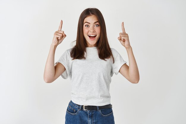 Überraschte und glückliche Frau, die Werbung zeigt, die Hände nach oben zeigt und fasziniert auf die Vorderseite schaut und über der weißen Wand steht.