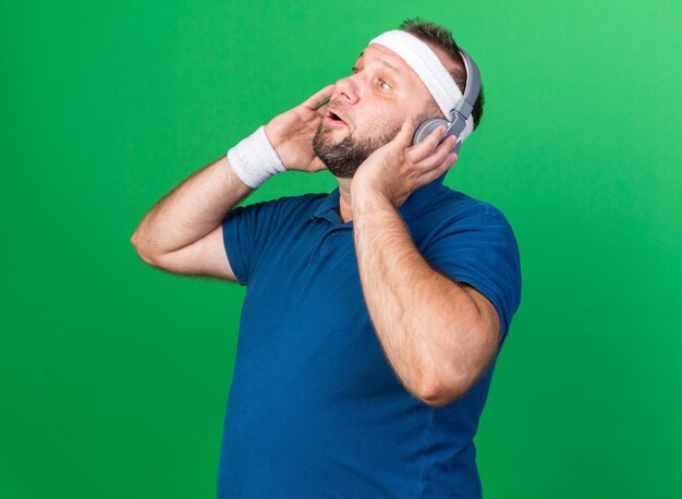 überraschter erwachsener slawischer sportlicher Mann mit Kopfhörern, der Stirnband und Armbänder trägt und seitlich isoliert auf grüner Wand mit Kopienraum schaut