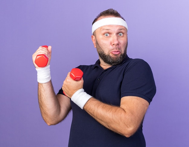 überraschter erwachsener slawischer sportlicher Mann mit Stirnband und Armbändern, die Hanteln isoliert auf lila Wand mit Kopierraum halten holding