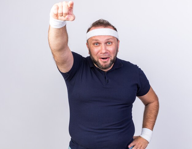 überraschter erwachsener slawischer sportlicher Mann mit Stirnband und Armbändern, die nach vorne zeigen, isoliert auf weißer Wand mit Kopierraum