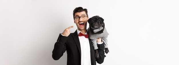 Kostenloses Foto Überraschter hundebesitzer, der auf seinen niedlichen schwarzen mops zeigt, der glücklichen welpen trägt, der weißen hintergrund trägt