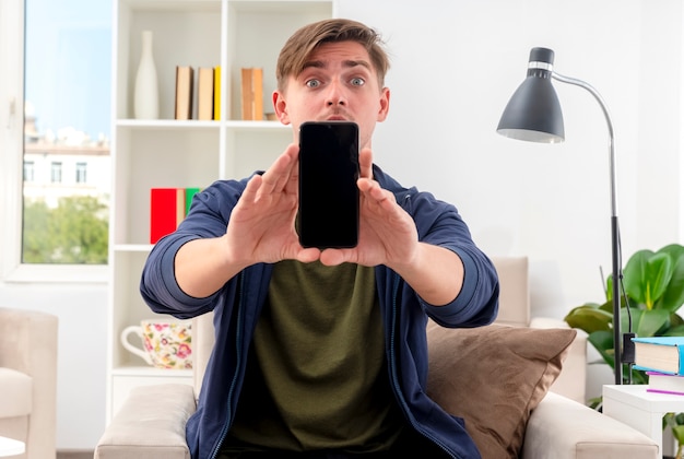 Überraschter junger blonder gutaussehender mann sitzt auf einem sessel und hält das telefon heraus Kostenlose Fotos