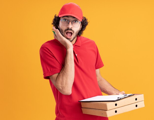 überraschter junger kaukasischer Lieferbote in roter Uniform und Mütze mit Brille, die Pizzapakete hält Klemmbrettstift mit Blick auf die Vorderseite und hält die Hand auf dem Gesicht