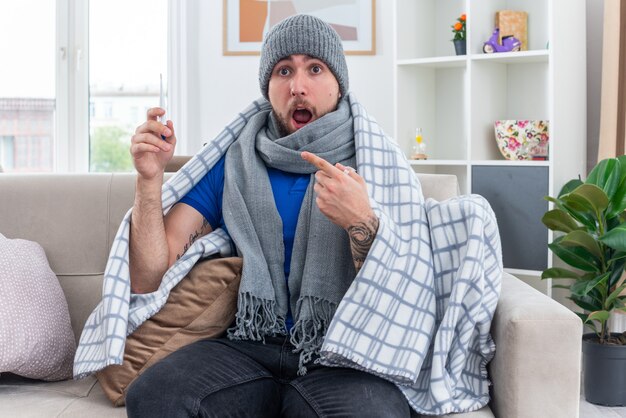 überraschter junger kranker Mann mit Schal und Wintermütze, der in Decke gehüllt auf dem Sofa im Wohnzimmer sitzt und auf das Thermometer zeigt, das nach vorne schaut