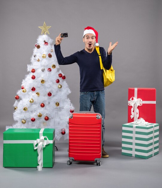 überraschter junger Mann, der Karte hält, die nahe Weihnachtsbaum steht und auf Grau präsentiert