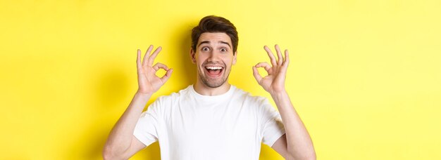 Überraschter junger Mann, der ok Zeichen zeigt und lächelt und etwas Gutes empfiehlt, das über gelbem Rücken steht