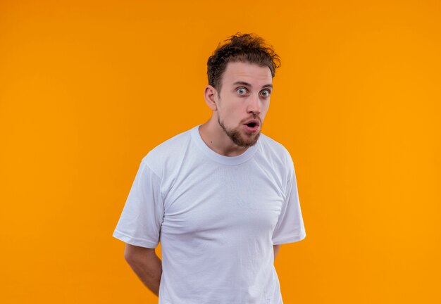 Überraschter junger Mann, der weißes T-Shirt hält Hände auf dem Rücken auf lokalisiertem orange Hintergrund hält