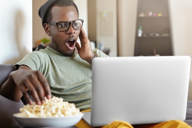 überraschter junger Mann mit Brille und Hut, der den Abend zu Hause mit einem Laptop verbringt