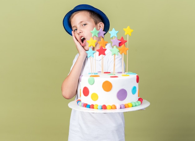 überraschter junger slawischer Junge mit blauem Partyhut, der Hand auf Gesicht legt und Geburtstagskuchen isoliert auf olivgrüner Wand mit Kopienraum hält