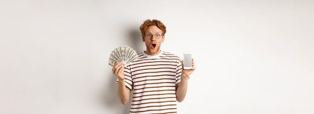 Kostenloses Foto Überraschter rothaariger mann, der smartphone-app auf leerem bildschirm zeigt und online-standi mit geldpreisen gewinnt