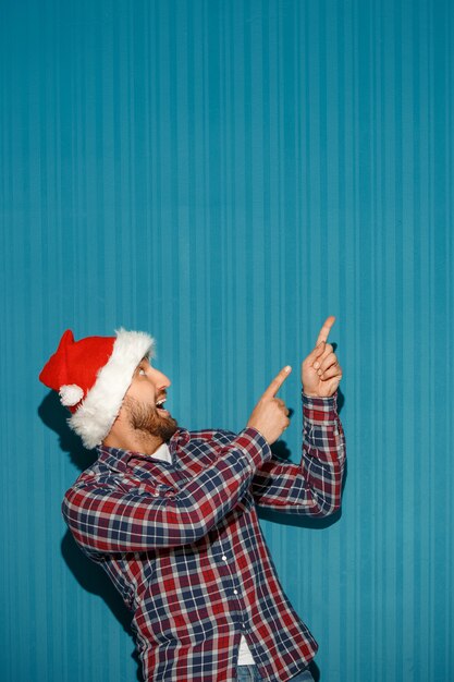 überraschter Weihnachtsmann, der eine Weihnachtsmütze trägt, die irgendetwas auf dem blauen Studiohintergrund zeigt