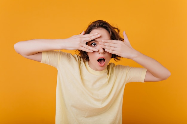 Kostenloses Foto Überraschtes dunkeläugiges mädchen mit lustigem ring, der auf orange wand aufwirft. blasse brünette frau im gelben t-shirt, das gesicht bedeckt und erstaunen ausdrückt.