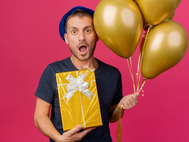 Überraschtes hübsches kaukasisches mann, das blauen parteihut trägt, hält heliumballons und geschenkbox lokalisiert auf rosa hintergrund mit kopienraum