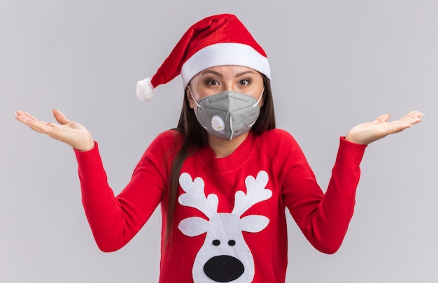 Kostenloses Foto Überraschtes junges asiatisches mädchen, das weihnachtsmütze mit pullover und medizinischer maske trägt, die hände lokalisiert auf weißem hintergrund verbreiten