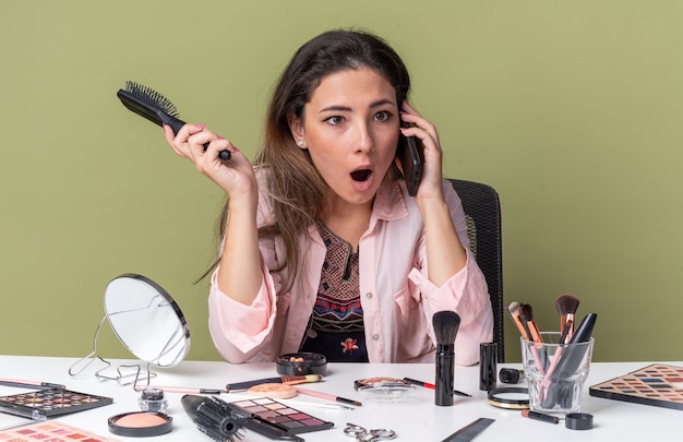 Überraschtes junges brünettes mädchen, das am tisch mit make-up-tools sitzt und am telefon spricht und den kamm isoliert auf olivgrüner wand mit kopienraum hält?