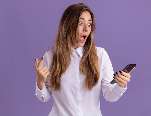 Überraschtes junges hübsches kaukasisches Mädchen hält und schaut auf das Telefon, das isoliert auf lila Wand mit Kopierraum zeigt